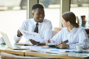 Cheerful handsome young black business expert showing diagram to female colleague while they analyzing sales and statistics in cafe