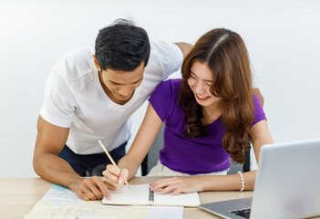 Asian happy female traveler sitting smiling at planning table using pencil taking note of tourist attractions in notebook while lovely husband standing helping searching information from paper map