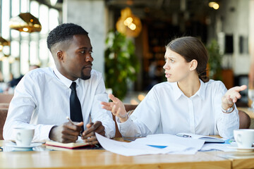 Puzzled young multi-ethnic business colleagues in white shirts sitting at table and discussing report data while working with papers in restaurant