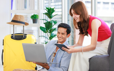 Obraz premium Asian young lovely boyfriend and girlfriend traveler couple sitting smiling on carpet in living room with trolley luggage while using laptop computer and tablet booking vacation summer trip online