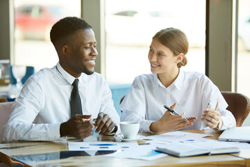 Positive young multi-ethnic business analysts sitting at table with papers and working with financial data while discussing development strategy
