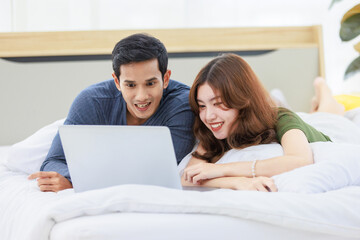 Fototapeta premium Asian happy romantic male female husband and wife lover couple lay down smiling together on pillow under blanket on bed using laptop notebook computer and credit card shopping making payment online