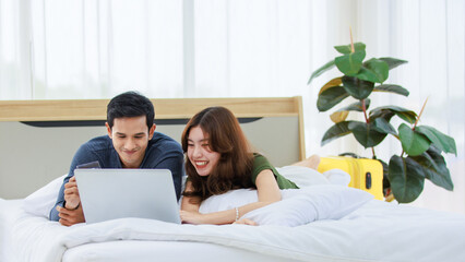 Obraz na płótnie Canvas Asian happy romantic male female husband and wife lover couple lay down smiling together on pillow under blanket on bed using laptop notebook computer and credit card shopping making payment online