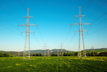 High voltage lines and power pylons in a flat and green agricultural mountain landscape on a sunny day with blue sky.
