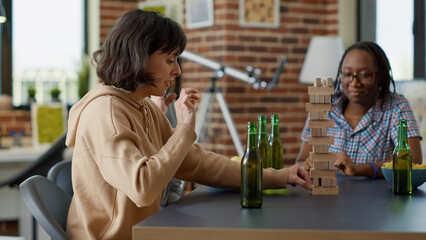 Young woman crashing building tower with wooden pieces on table, losing board game competition. Person and happy friends playing games with square blocks and cubes at fun gathering.