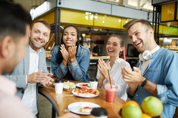Group of positive young multi-ethnic people in casual outfits sitting at table with snacks and applauding while congratulating friend in cafe