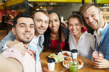 Smiling handsome young Latin man with stubble taking selfie with friends in modern cafe