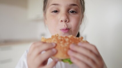 little kid girl eating a hamburger. unhealthy fast food meal proper lifestyle nutrition concept. child greedily with pleasure bites a big burger in the kitchen at home. kid eats fast food close-up