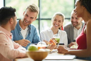 Cheerful excited young multi-ethnic friends in casual outfits standing at table and eating snacks while laughing together