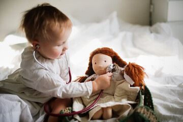 Cute girl child toddler listens with stethoscope to a doll toy in the bed in the bedroom. The child...