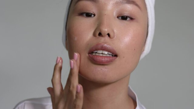 Closeup of asian woman with towel on head applying white cream on face. Neutral natural pink manicure on nails. Long fingers. Beautiful face with big lips. Woman doing cosmetic procedure, preparing.