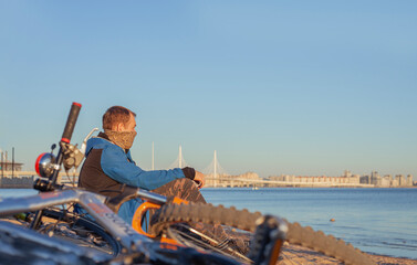 Man in jacket with snood and bicycle sitting in sand on beach by sea. Cyclist with face mask against dust. Cycling in city in spring or autumn. Man sitting next to mountain bikes with cityscape.