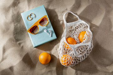 leisure and summer holidays concept - string bag of oranges, earbuds, book and sunglasses on beach...