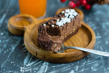Appetizing slice of chocolate brownie in a spoon, brownie on a wooden plate, glass of multifruit juice and new year's decor on the background