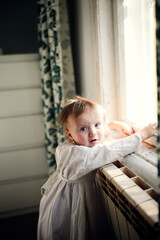 Cute baby toddler girl in nightgown at the window in the bedroom, close-up portrait
