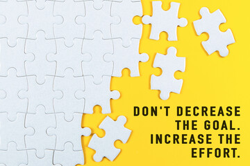 Don't decrease the goal. Increase the effort. Motivational text on white jigsaw puzzle with some missing pieces on yellow background.