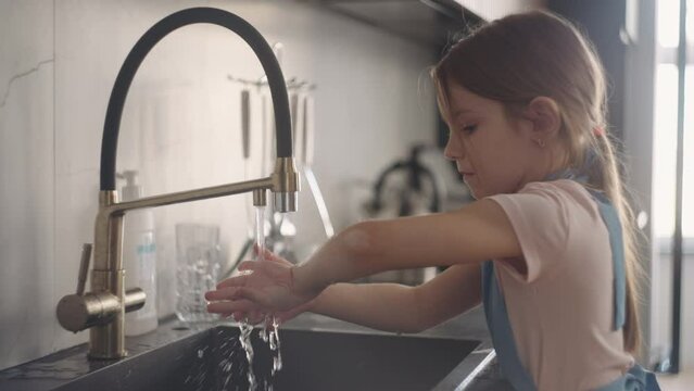 washing hands in kitchen, little girl in apron is rubbing her palms under water stream from tap