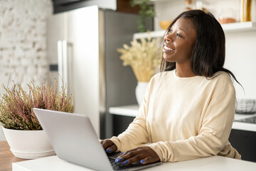 Inspired African-American freelancer woman enjoying remote work from home, cheerful lady using laptop sitting in the kitchen at home, looks away dreamly