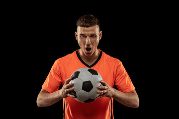 Front camera view of young male soccer player in orange-blue football kit posing with ball isolated on dark background. Concept of sport, goals, competition, emotions © master1305