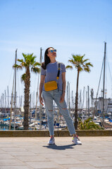 Young 30 years old woman in jeans, t-shirt and yellow bag walking in Barcelona harbor or port or...