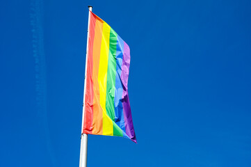 LGBTIQ flag waving on the wind on flagpole in town. A symbol of the LGBT community and social movements. LGBTIQ flag against blue sky. Symbol of lesbian, gay, bisexual, transgender and queer community