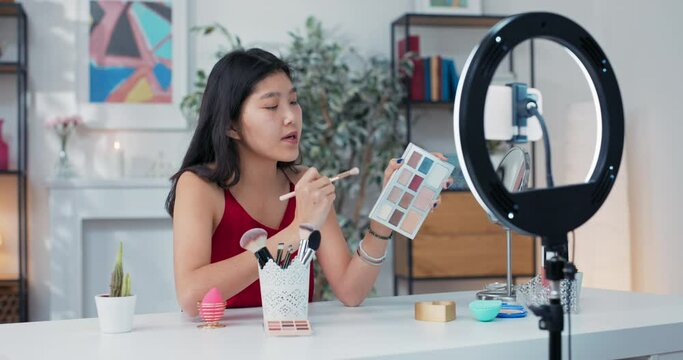 Experienced makeup artist of asian descent applies eye shadow on the eyelids, recording a tutorial on smoky eyes, in front of her on the table mirror, cosmetics and beauty accessories.
