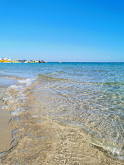 A small wave runs on the sandy shore of the beach in Fig Tree Bay against the background of the clear water of the Mediterranean Sea and the blue cloudless sky.