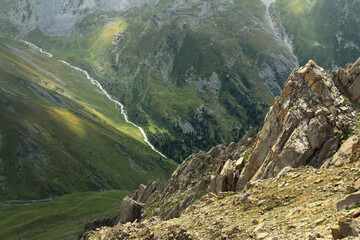 Sharp rocks on the slope of the mountain with a distant view of the alpine valley of the Prokhodnoye gorge, a river flows through the gorge, sunlight on the slopes, view from above