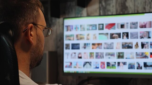 Close-up of a bearded man wearing glasses at night watching erotic photos on the monitor screen. Sexual addiction. Sex alone. Male masturbation