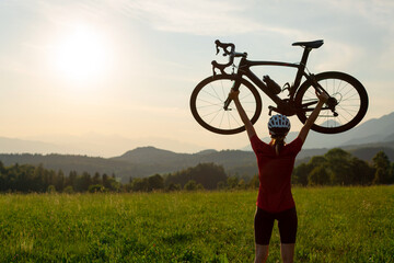 Naklejka premium Female road cyclist raising bicycle with both arms above her head at sunset with beautiful green nature. Winning, success and achievement concept.