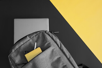 Gray textile urban backpack with a laptop, notepad and fountain pen on a black and yellow table. Study and lifestyle with gadgets. Copy space.