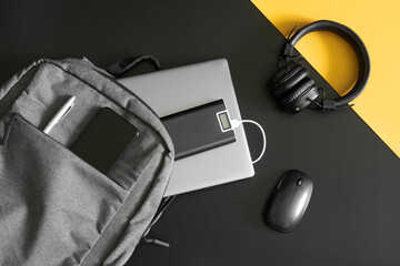 Gray textile urban backpack with laptop, external battery, phone, headphones, mouse and pen on black and yellow background. Travel, trip, study and lifestyle with gadgets. Recharging devices. - obrazy, fototapety, plakaty