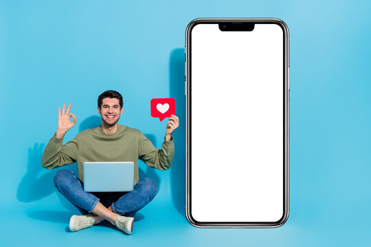 Full length portrait of positive man sit floor show okey symbol huge phone isolated on blue color background