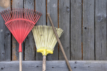 A fan rake and a whisk stand upright against the plank wall of the barn. Rustic tools near the barn...