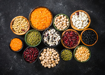 Legumes, lentils, chikpea and beans assortment in different bowls on black stone table. Top view.