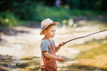 Preschooler boy in yellow rubber boots plays fishing, child fishes with stick near lake in countryside, games in nature and childhood without gadgets, summer holidays