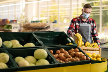 Caucasian handsome man worker in mask and apron walking in supermarket with basket of fruits. Male young food store assistant working in retail. Job