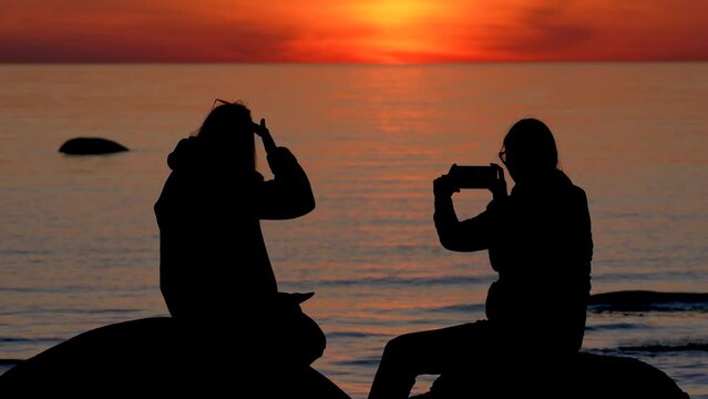Young women silhouettes sitting by water during beautiful sunset. Friends talking, gesturing, taking photo and video using smartphone.