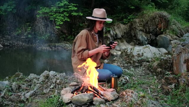 Relaxation young caucasian woman in hat holding smartphone sitting near calm bonfire and wild river against backdrop of nature at evening time. Local travel, tourism, camping and blogger concept