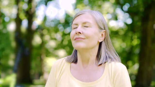 Close up portrait senior retiree woman standing in nature between forest trees relaxes, breathes fresh air closing eyes. Mature female enjoys a life of peace calm, quiet in the park. Outdoor Happiness