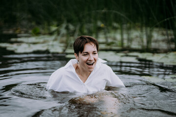 A young woman with a short haircut swims in a forest lake and laughs. Portrait