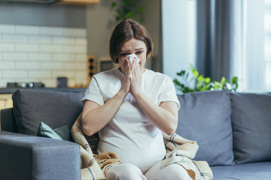 Sad pregnant woman at home sitting on the couch and crying, has a runny nose and allergies sneezes into a napkin