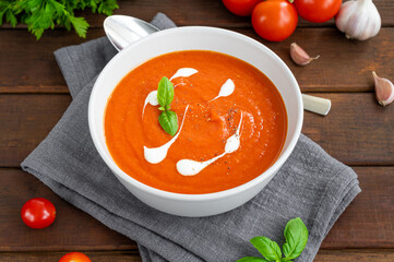 Tomato soup puree in a bowl with cream and fresh basil leaves on a dark wooden background. Copy space. Top view.