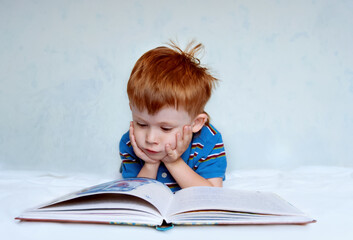 A cute little boy with red hair and a blue shirt is lying on the bed reading a book