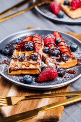Belgian waffle with chocolate, strawberry, blueberries and powdered sugar on dark plate