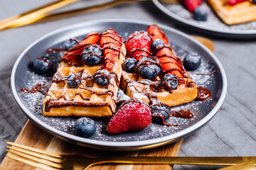 Belgian waffle with chocolate, strawberry, blueberries and powdered sugar on dark plate