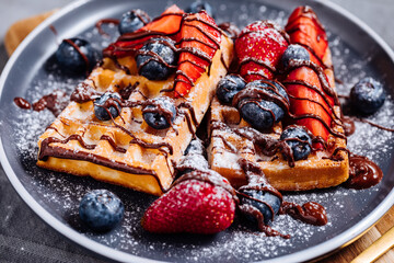 Belgian waffle with chocolate, strawberry, blueberries and powdered sugar on dark plate