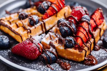 Belgian waffle with chocolate, strawberry, blueberries and powdered sugar on dark plate