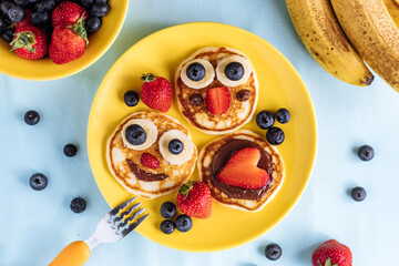 American pancakes decorated like smile and happy faces with strawberries, chocolate, blueberries and banana. Food for kids, playful and creative.
