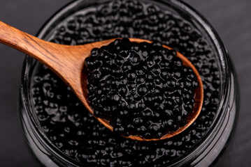 Wooden spoon with black caviar against the background of black caviar in a jar.